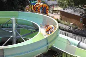 Große grüne Wasserrutsche auf einem Campingplatz, daneben Gebäude, Zaun und Bäume im Hintergrund.