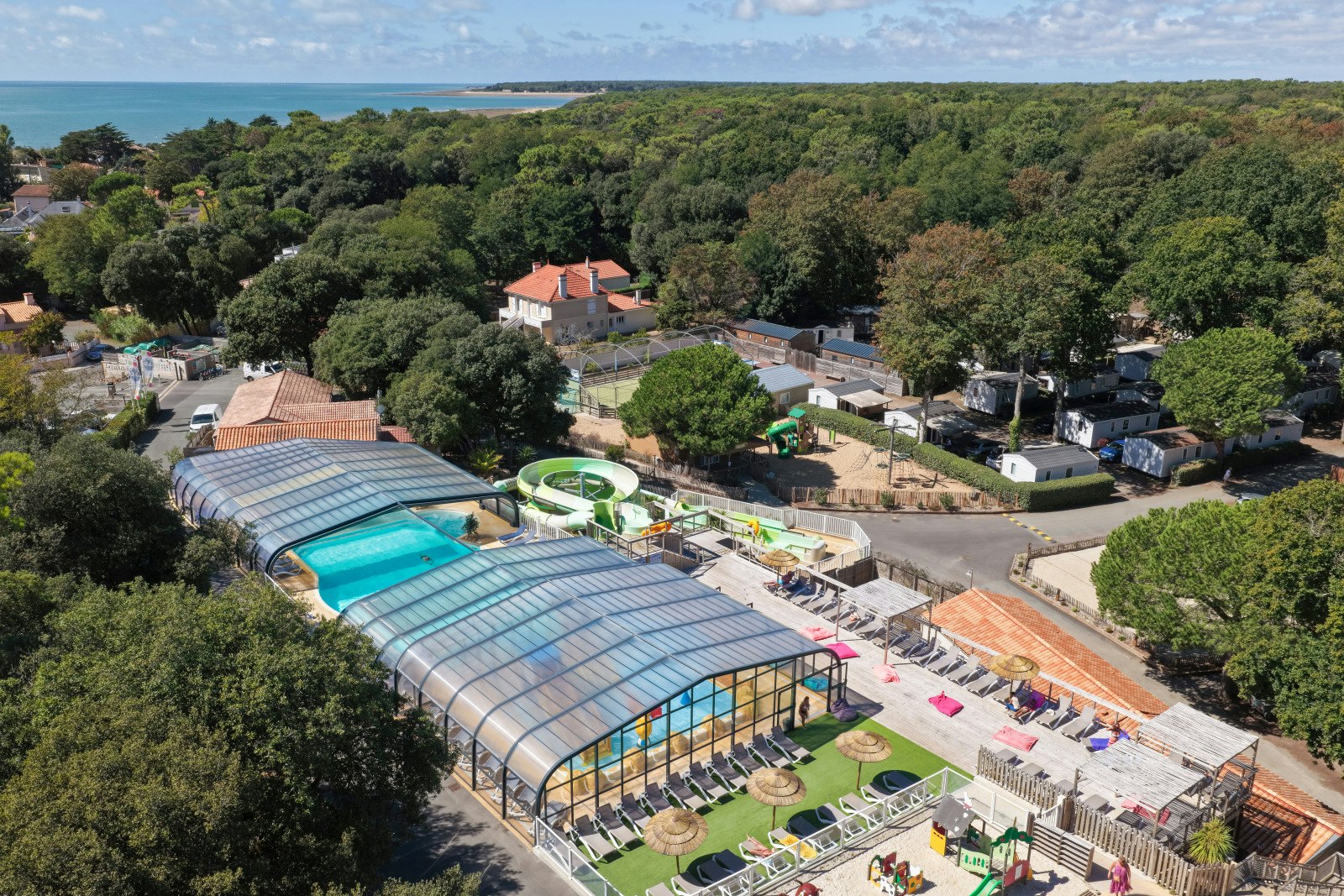 Luftaufnahme eines Campingplatzes mit beheiztem Schwimmbecken, Glasdach-Halle, Stellplätzen und Wegen zwischen Bäumen und Häusern.