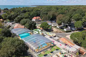 Luftaufnahme eines Campingplatzes mit beheiztem Schwimmbecken, Glasdach-Halle, Stellplätzen und Wegen zwischen Bäumen und Häusern.