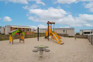 Spielplatz am Campingplatz mit gelber Rutsche, Federwippe und Sandfläche, umgeben von Wohnwagen und Gartenzaun.
