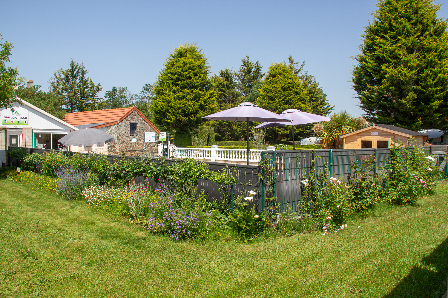 Gepflegter Garten mit Blumenbeeten, Zaun und Terrasse mit Sonnenschirm vor Ferienhäusern und Bäumen.