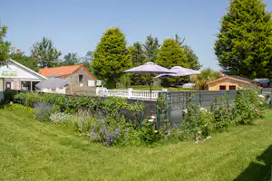 Gepflegter Garten mit Blumenbeeten, Zaun und Terrasse mit Sonnenschirm vor Ferienhäusern und Bäumen.
