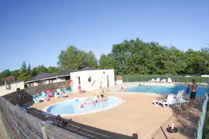 Camping-Pool mit Kinderbecken, Sonnenliegen und Spielzeug im Wasser, im Hintergrund ein weißes Gebäude und Bäume.