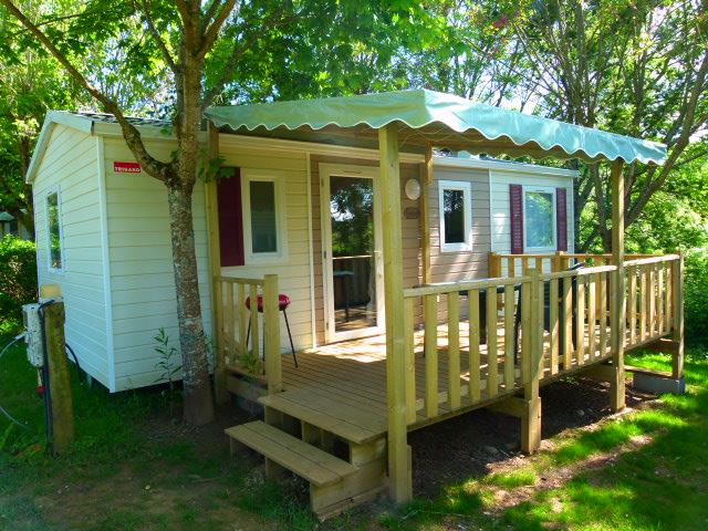 Bungalow mit Holzveranda und Markise, seitlich ein weiteres Wohnmodul, umgeben von Bäumen und Rasen.