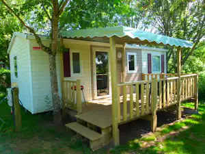 Bungalow mit Holzveranda und Markise, seitlich ein weiteres Wohnmodul, umgeben von Bäumen und Rasen.