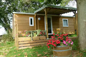 Holz-Bungalow mit Veranda und Sitzbereich, Blumen im Kübel vor dem Eingang, umgeben von Bäumen und Rasen.