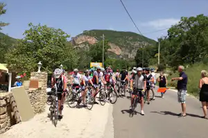 Radfahrergruppe auf einer Straße am Campingplatz, Fahrräder nebeneinander, Steinmauer, Bäume und Felsberg im Hintergrund.