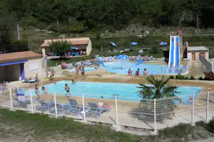 Schwimmbecken mit Wasserrutsche, Liegestühlen und Sonnenschirmen auf dem Campingplatz, im Hintergrund Bungalows und Bäume.