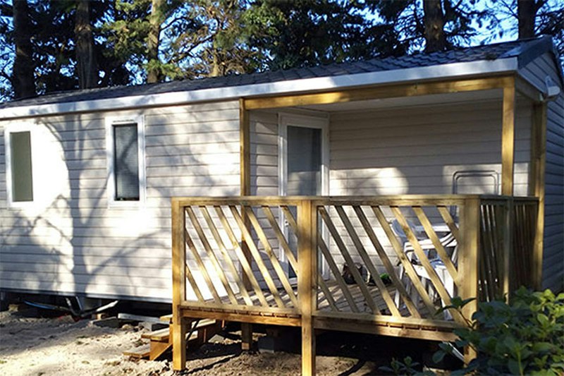 Wohnwagen mit Holzterrasse und Stufen, umgeben von Büschen und Bäumen, im Campingplatzbereich.