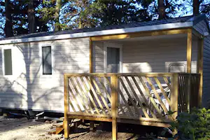 Wohnwagen mit Holzterrasse und Stufen, umgeben von Büschen und Bäumen, im Campingplatzbereich.