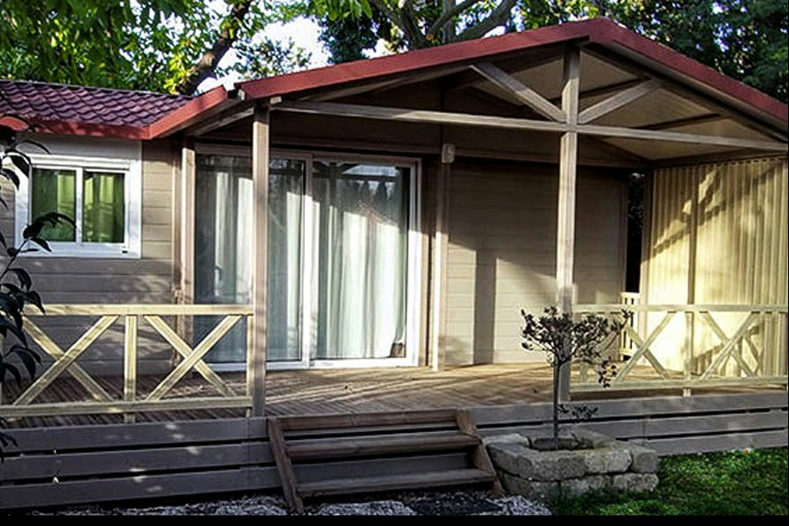 Bungalow mit überdachter Terrasse, großen Fenstern und Holzgeländer, davor Stufen und ein kleiner Steinweg im Garten.