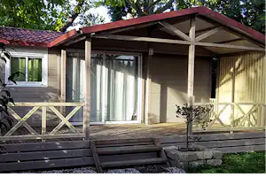 Bungalow mit überdachter Terrasse, großen Fenstern und Holzgeländer, davor Stufen und ein kleiner Steinweg im Garten.