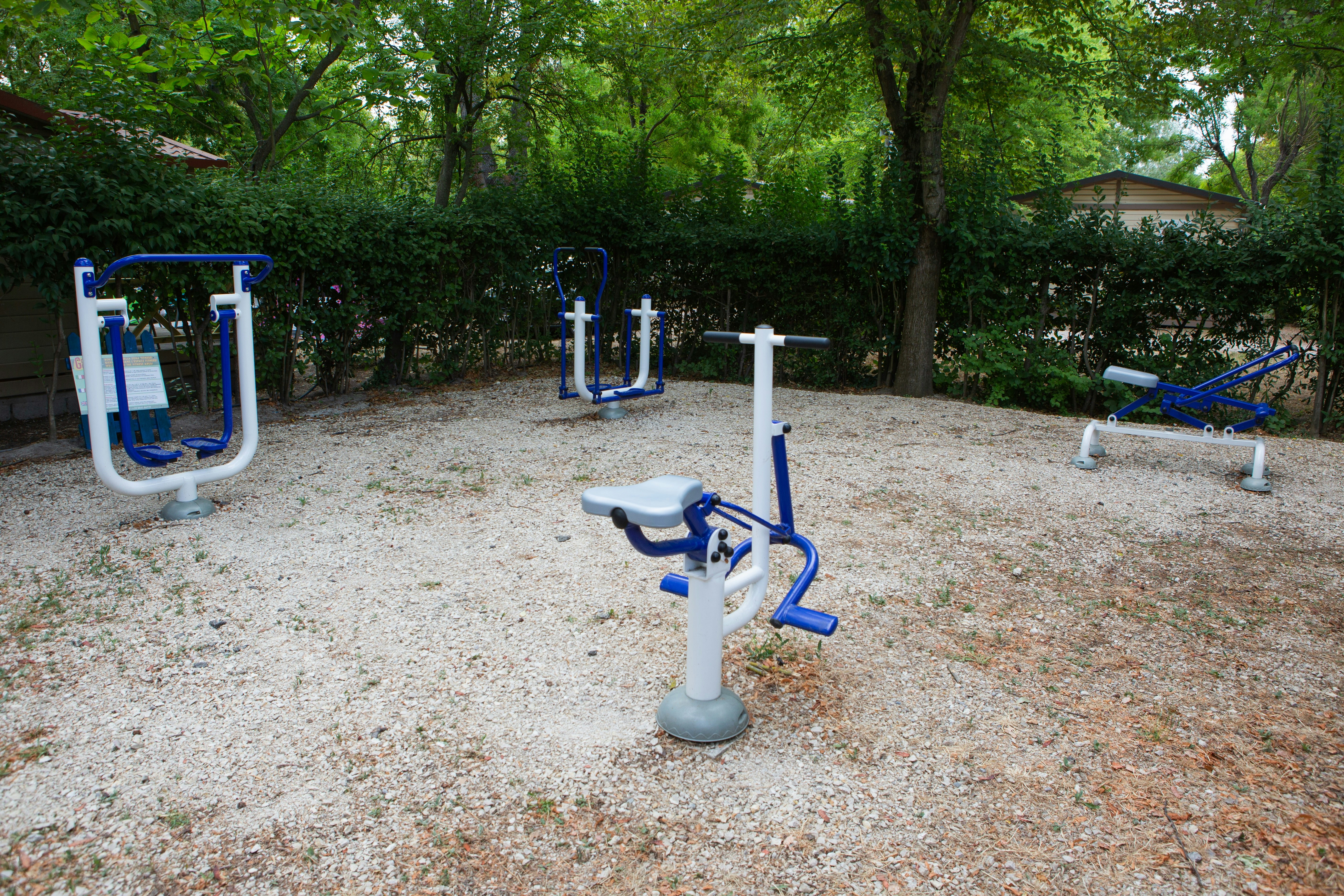 Fitnessgeräte auf einem Kiesplatz im Campingbereich, umgeben von Bäumen und Hecken.