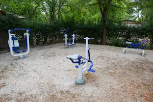 Fitnessgeräte auf einem Kiesplatz im Campingbereich, umgeben von Bäumen und Hecken.