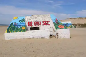 Bunt bemalte Mauer mit Schriftzug „LE AIN SEC“ am Sandstrand, dahinter Dünen und das Meer.