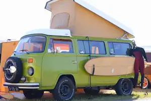 Grüner Camper-Van mit Dachzelt und geöffnetem Vorzelt, am Stellplatz vor einem Wohnwagen-Anhänger.