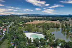 Luftaufnahme vom Campingplatz mit Schwimmbecken, kleinem See, Bäumen und Feldern in der Landschaft.