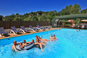 Familie im Pool mit Schwimmreifen, Holzdeck und Liegestühlen am Campingplatz, dahinter Zaun und Bäume.