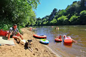 Mann sitzt am Seeufer mit zwei Hunden, daneben liegen rote und grüne Schlauchboote im Wasser.