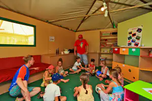 Kinder spielen in einem Indoor-Spielraum mit grünem Boden, roten Sitzbänken und einem Betreuer unter einer Zeltdecke.