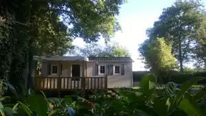 Mobilheim mit Veranda und Holzgeländer, umgeben von Büschen und Bäumen, im Campingbereich.