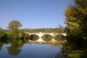 Steinbogen-Brücke über einem ruhigen Fluss, umgeben von Bäumen und grünem Ufer, im klaren Himmel.