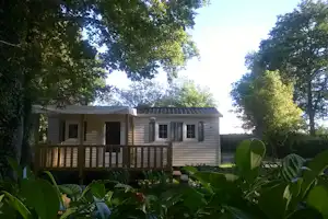 Mobilheim mit Veranda und Markise, umgeben von Bäumen und Pflanzen im Campingbereich.