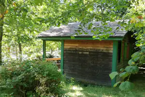 Holzhütte mit Satteldach im Grünen, umgeben von Bäumen und Sträuchern, mit Grasfläche davor.