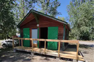Holzbungalow mit grünen Türen und Veranda auf Holzpodest, umgeben von Bäumen und Kiesboden.