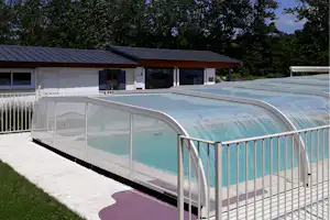 Überdachter Pool mit Glaswand und Geländer vor einem weißen Gebäude, im Hintergrund Bäume und grünes Gelände.