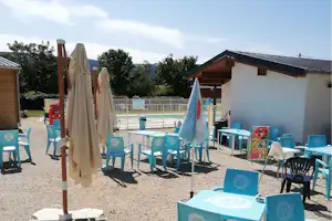 Terrasse am Campingplatz mit blauen Tischen und Stühlen, Sonnenschirmen, Pool im Hintergrund und Gebäude am Rand.