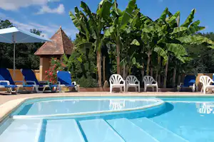 Camping-Pool mit Liegestühlen, Sonnenschirm und Gartenhaus im Hintergrund, dazu Bananenstauden und Blumenbeet.