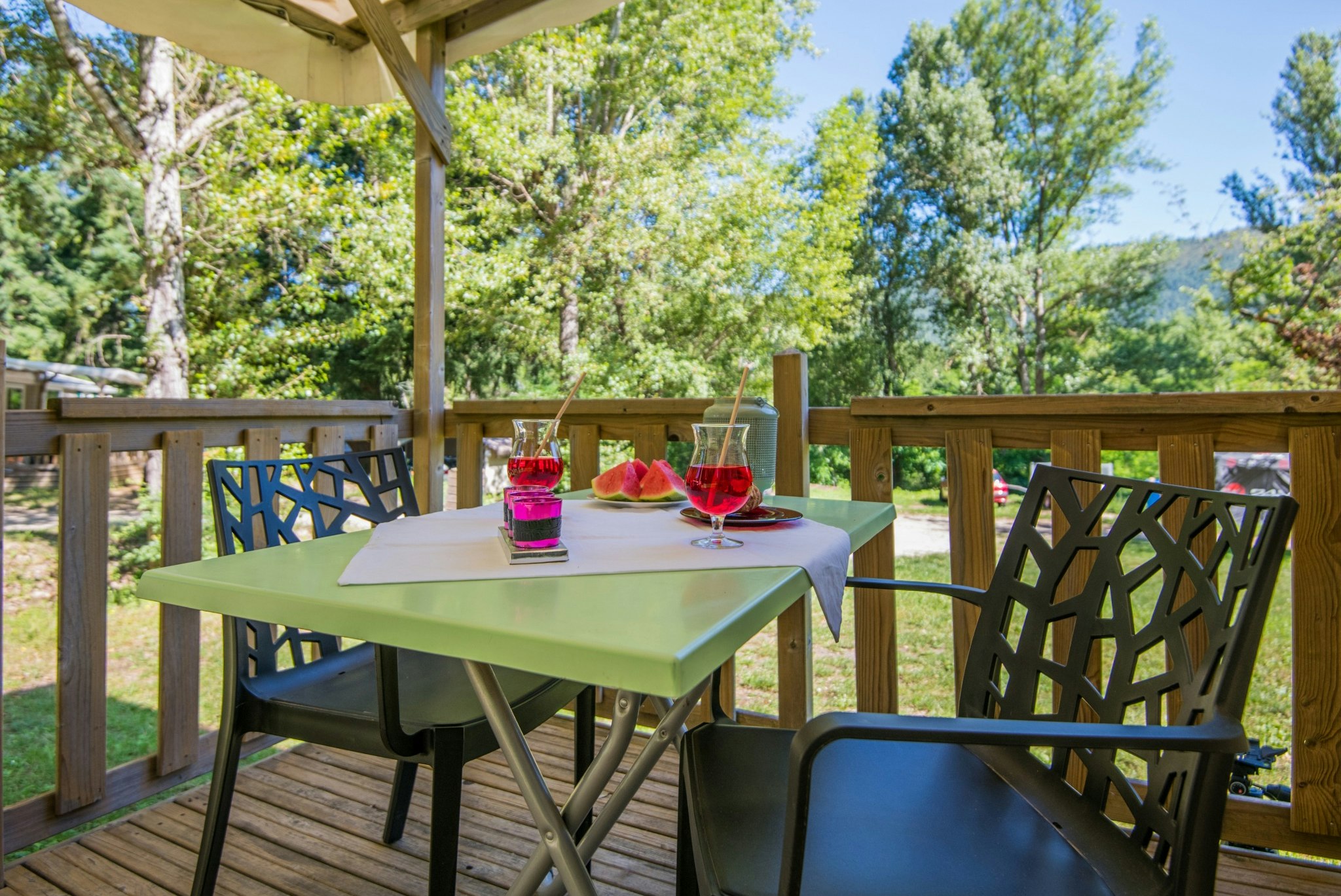 Grüner Tisch auf der Holzterrasse mit Gläsern und Wassermelone, Sonnenschirm und Blick in den Garten mit Bäumen.