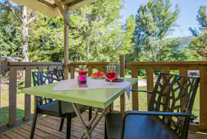 Grüner Tisch auf der Holzterrasse mit Gläsern und Wassermelone, Sonnenschirm und Blick in den Garten mit Bäumen.