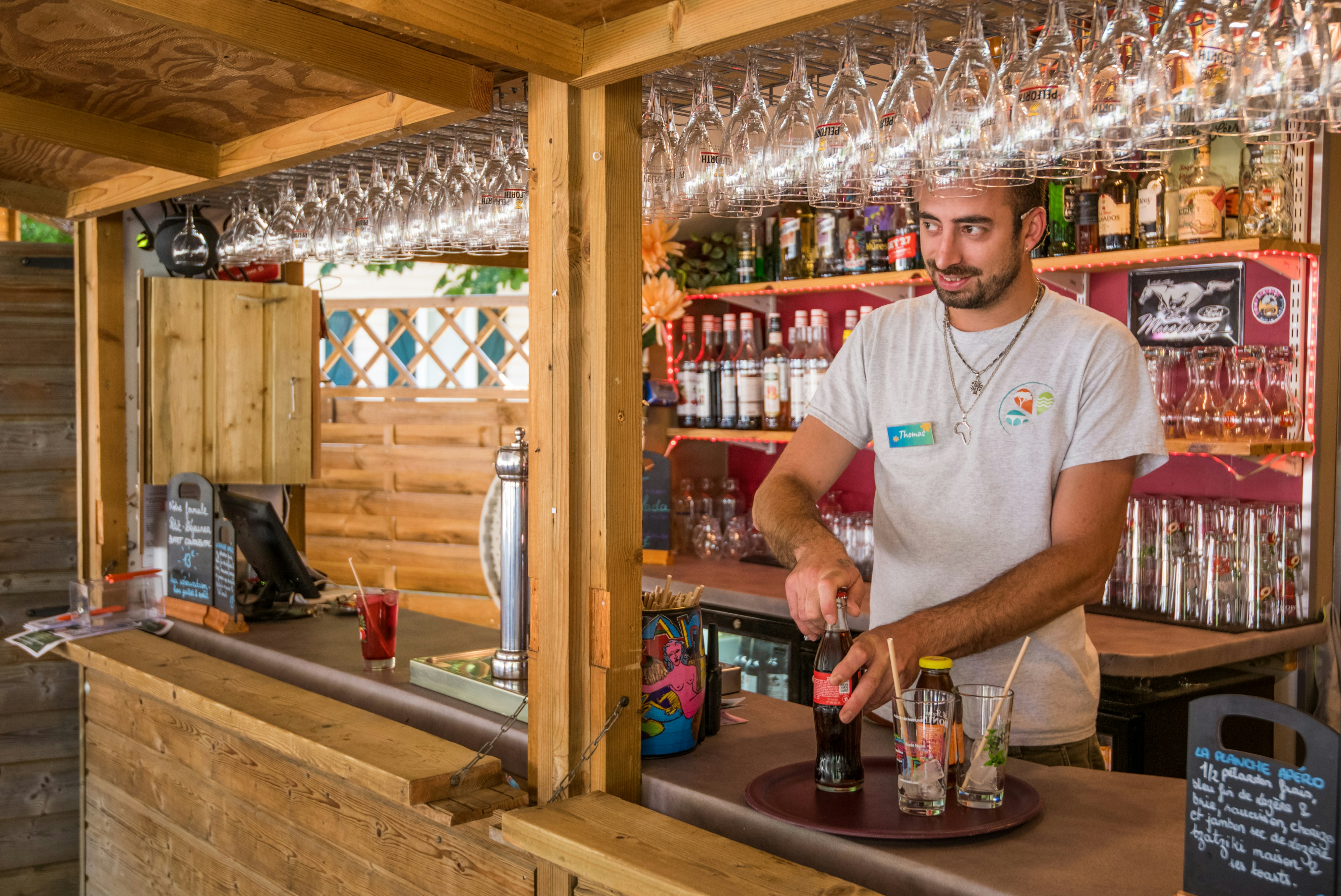 Campingbar mit Holztheke, vielen Biergläsern und Flaschen; ein Mitarbeiter schenkt ein.