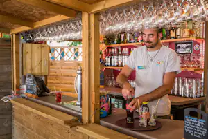 Campingbar mit Holztheke, vielen Biergläsern und Flaschen; ein Mitarbeiter schenkt ein.