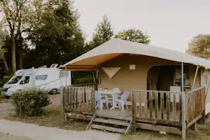 Campingzelt mit Holzterrasse, Tisch und Stühlen; daneben ein weißer Van am Stellplatz im Grünen.