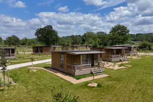 Holzunterkünfte auf einem Campingplatz mit Wegen, Kiesflächen und Sitzbereichen, umgeben von Bäumen und Hügeln.