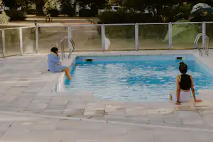 Campingpool mit blauem Wasser, Edelstahlleiter und Sichtschutzzaun; zwei Personen sitzen am Beckenrand auf Steinfliesen.