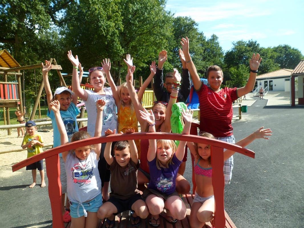 Kinder auf einem Spielplatz im Campingbereich, mit Kletterbogen und bunten Spielgeräten vor Wohnwagen und Bäumen.