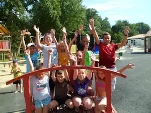 Kinder auf einem Spielplatz im Campingbereich, mit Kletterbogen und bunten Spielgeräten vor Wohnwagen und Bäumen.