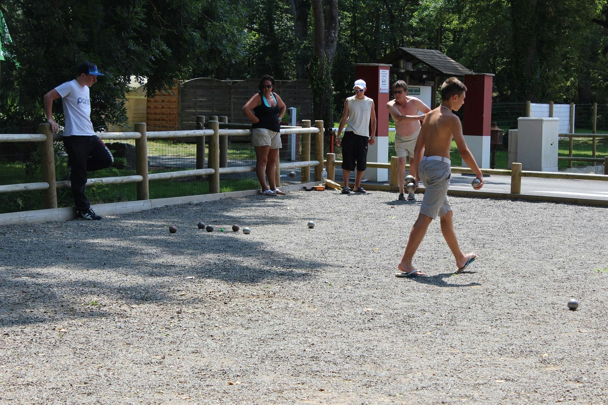 Boule-Spiel auf dem Campingplatz: mehrere Personen stehen auf dem Kiesplatz, im Hintergrund Bäume und ein Gebäude.