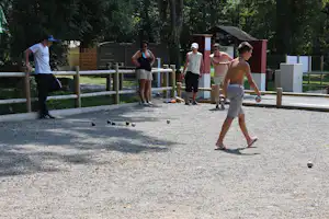 Boule-Spiel auf dem Campingplatz: mehrere Personen stehen auf dem Kiesplatz, im Hintergrund Bäume und ein Gebäude.
