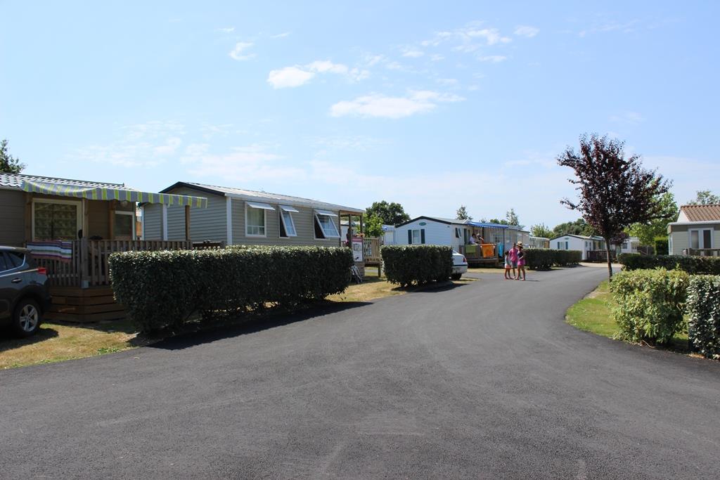 Reihe von Wohnwagen und Mobilheimen auf einem Campingplatz, mit gepflastertem Weg, Hecken und geparktem Auto.