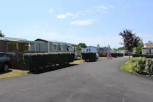 Reihe von Wohnwagen und Mobilheimen auf einem Campingplatz, mit gepflastertem Weg, Hecken und geparktem Auto.