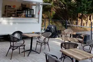 Wohnwagen mit überdachter Terrasse, Holztisch und Stühlen sowie Gartenmöbeln, im Hintergrund Zaun und Bäume.