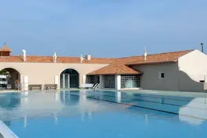 Schwimmbecken am Campingplatz mit Pooltreppe, Liegen und weißem Gebäude mit Terrassenbögen und Ziegeldach.