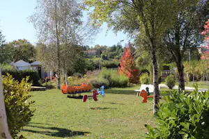 Spielplatz im Campinggarten mit bunten Tierwippen, Rasenfläche, Bäumen und Hecken, im Hintergrund Zelte und Wohnwagen.