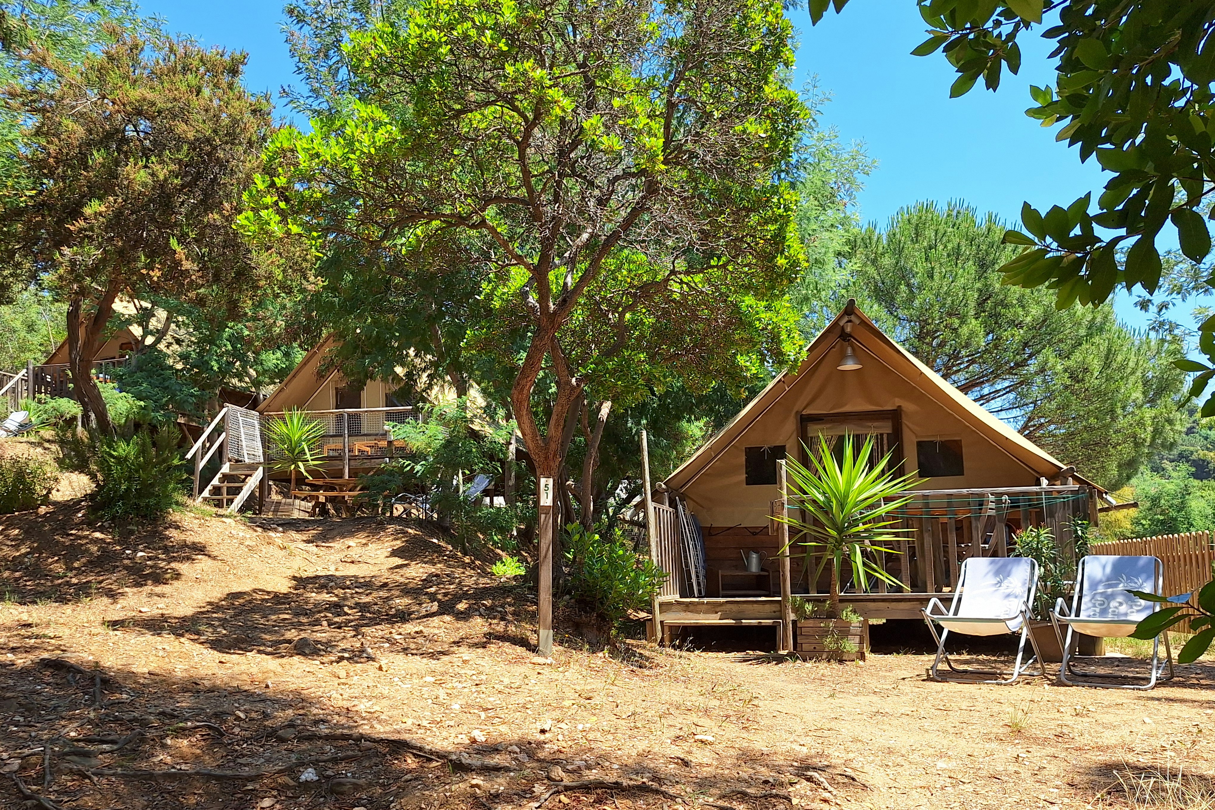 Holz-Zelt mit Veranda und Stühlen im Campingbereich, umgeben von Bäumen und Wegen, im Hintergrund weitere Unterkünfte.