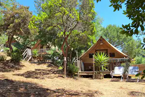 Holz-Zelt mit Veranda und Stühlen im Campingbereich, umgeben von Bäumen und Wegen, im Hintergrund weitere Unterkünfte.